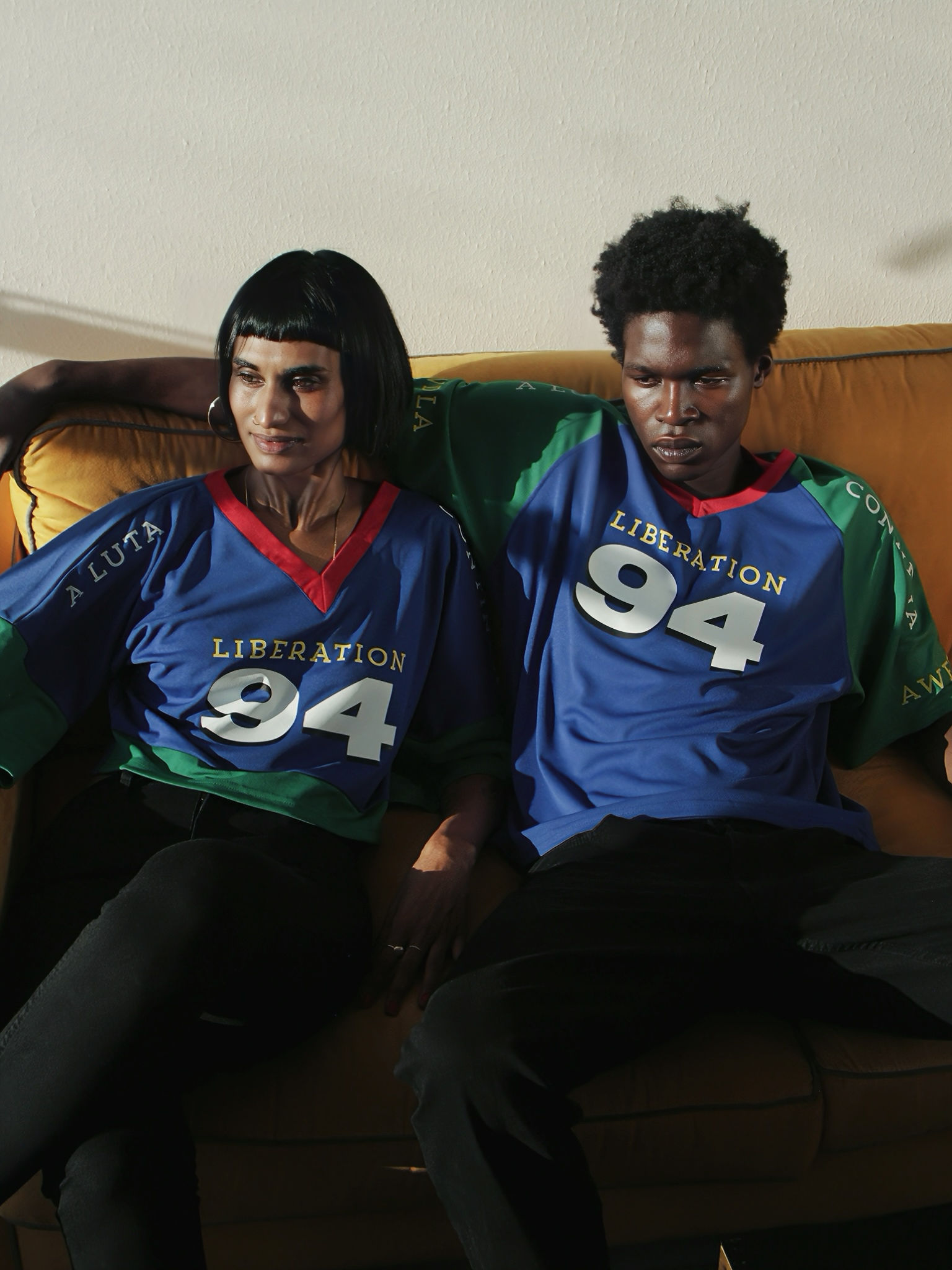 Two models wearing blue and green Liberation 94 shirts seated on a couch.